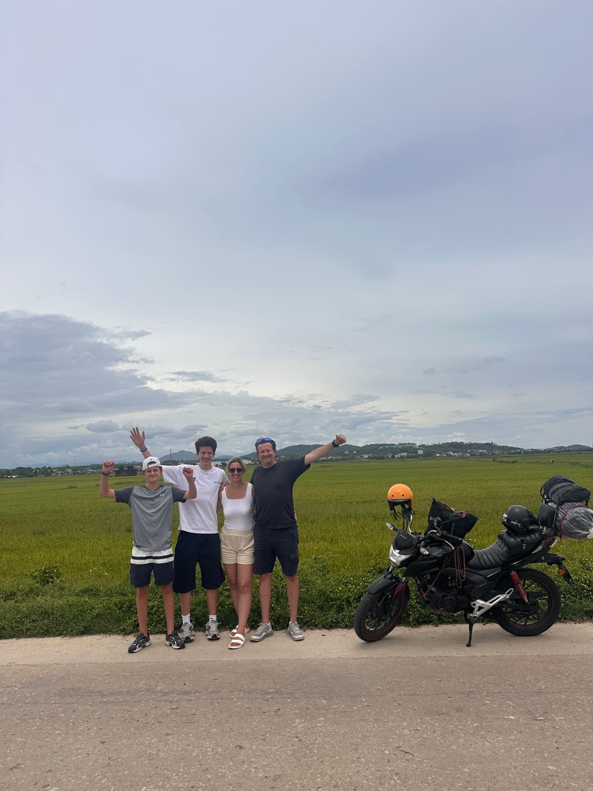 A family of four posing arms-up beside a loaded motorbike and rice fields