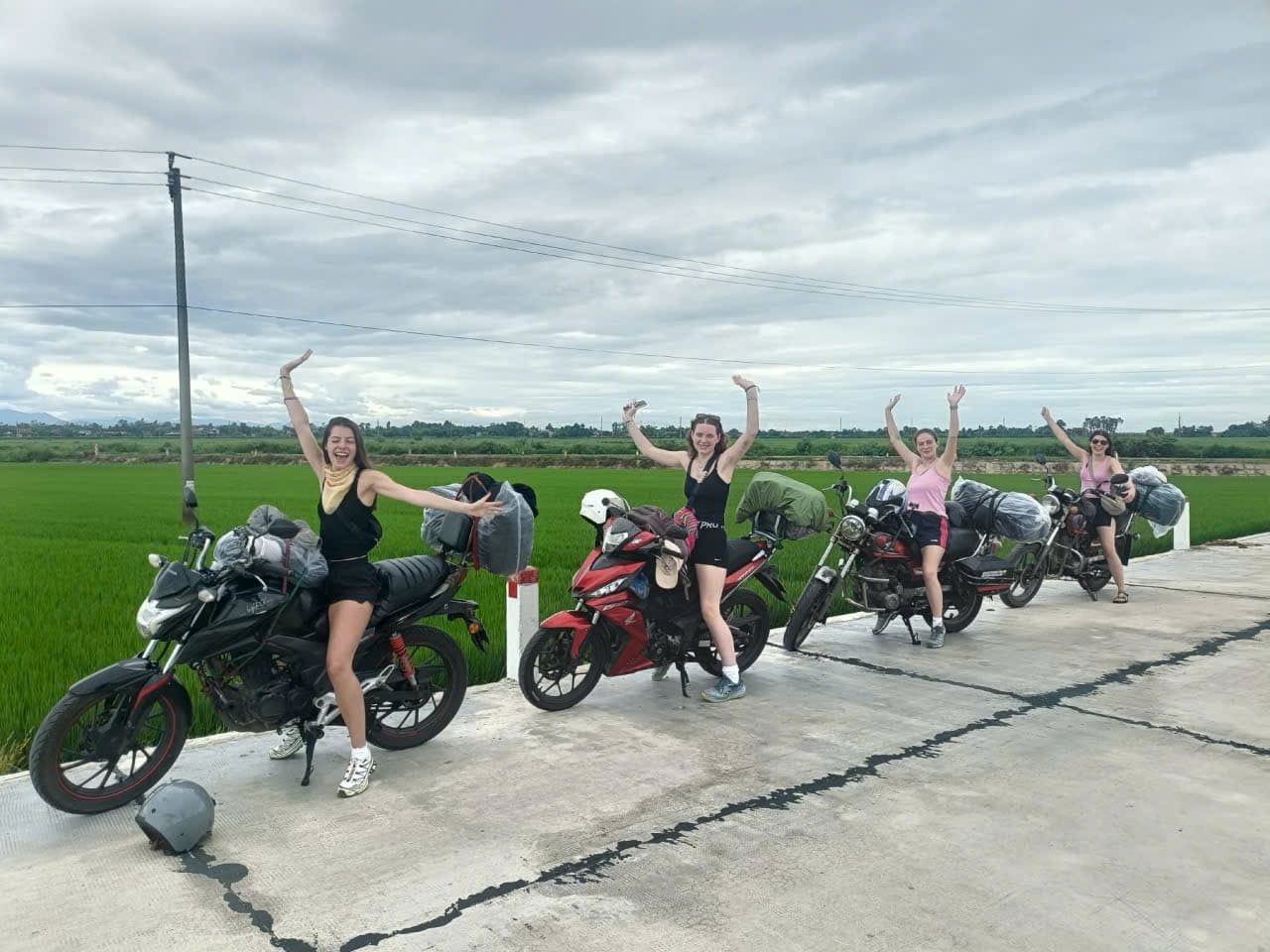 Four guests posing with motorbikes and strapped luggage beside a bright green rice paddy